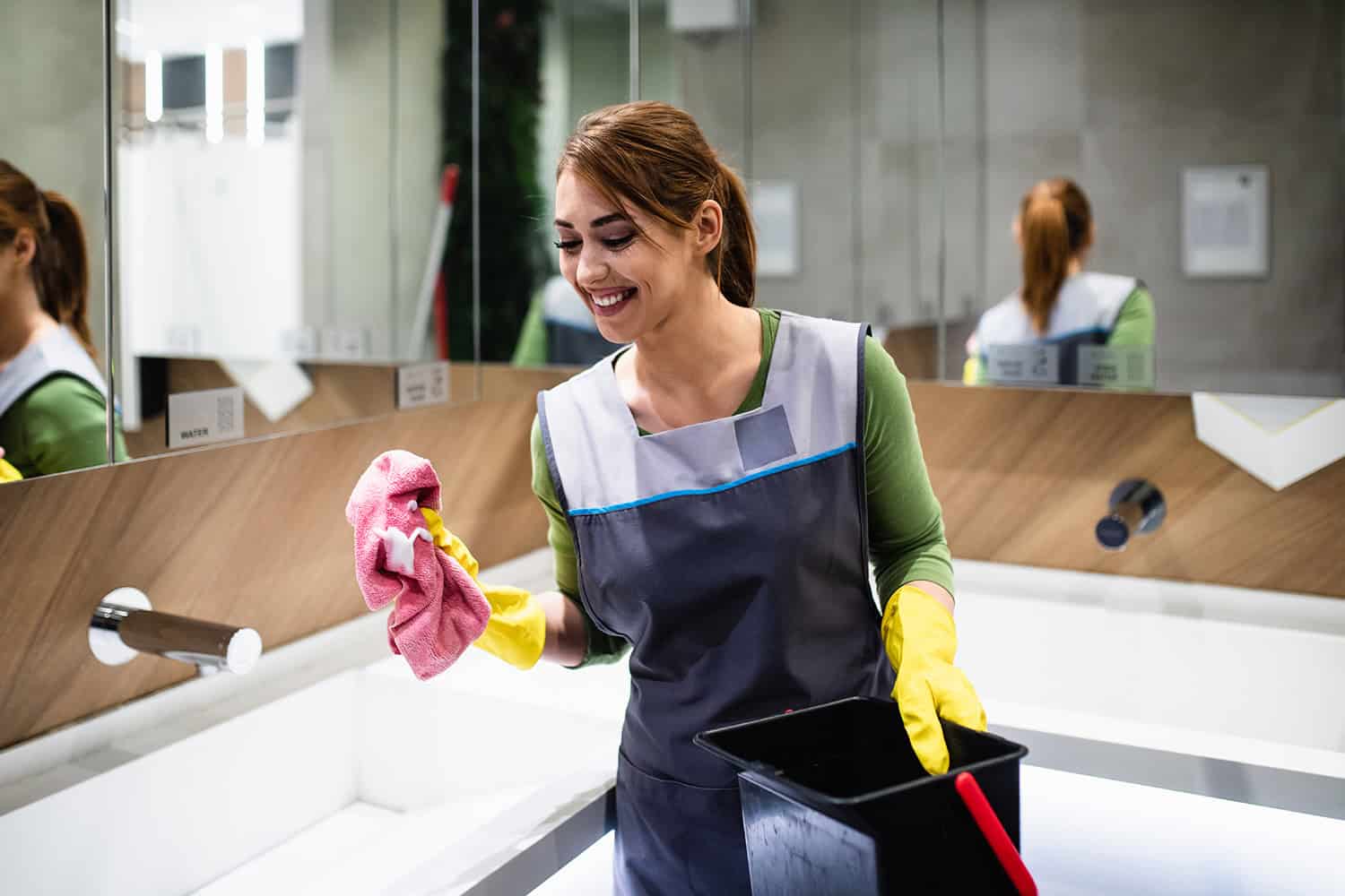 Cleaner wearing yellow gloves cleaning shopping centre bathroom with pink cloth